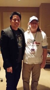 Two men posing together indoors, one in a blazer and the other in casual attire with a cap.