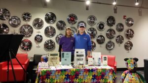 Two women standing behind a colorful table with tech gadgets at an event.