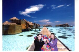 Two women posing happily on a wooden walkway over clear blue water under a sunny sky.