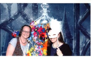 Two people posing with a colorful puzzle-piece outfit and a white feathered mask.