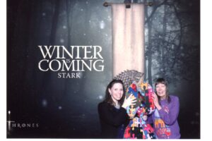 Two women posing with a colorful dragon prop under a 'Winter is Coming' Stark banner.