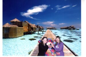 Two women posing on a wooden pier with overwater bungalows and clear blue sky.