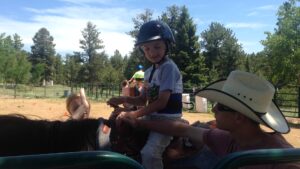 A young child wearing a helmet sits on a horse with adult assistance outdoors.