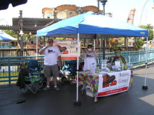 Two people stand under a blue canopy at a community event booth.