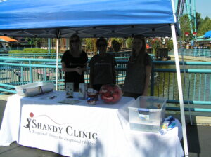 Three people stand behind a Shandy Clinic booth under a blue canopy.