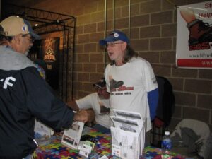 Two men shaking hands at an indoor event table with brochures and items.