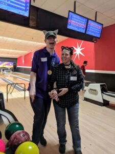Two men smiling together at a bowling alley, one wearing a cat hat.