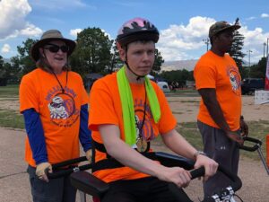 Three people in orange shirts outdoors, one in a wheelchair with a helmet and medals.